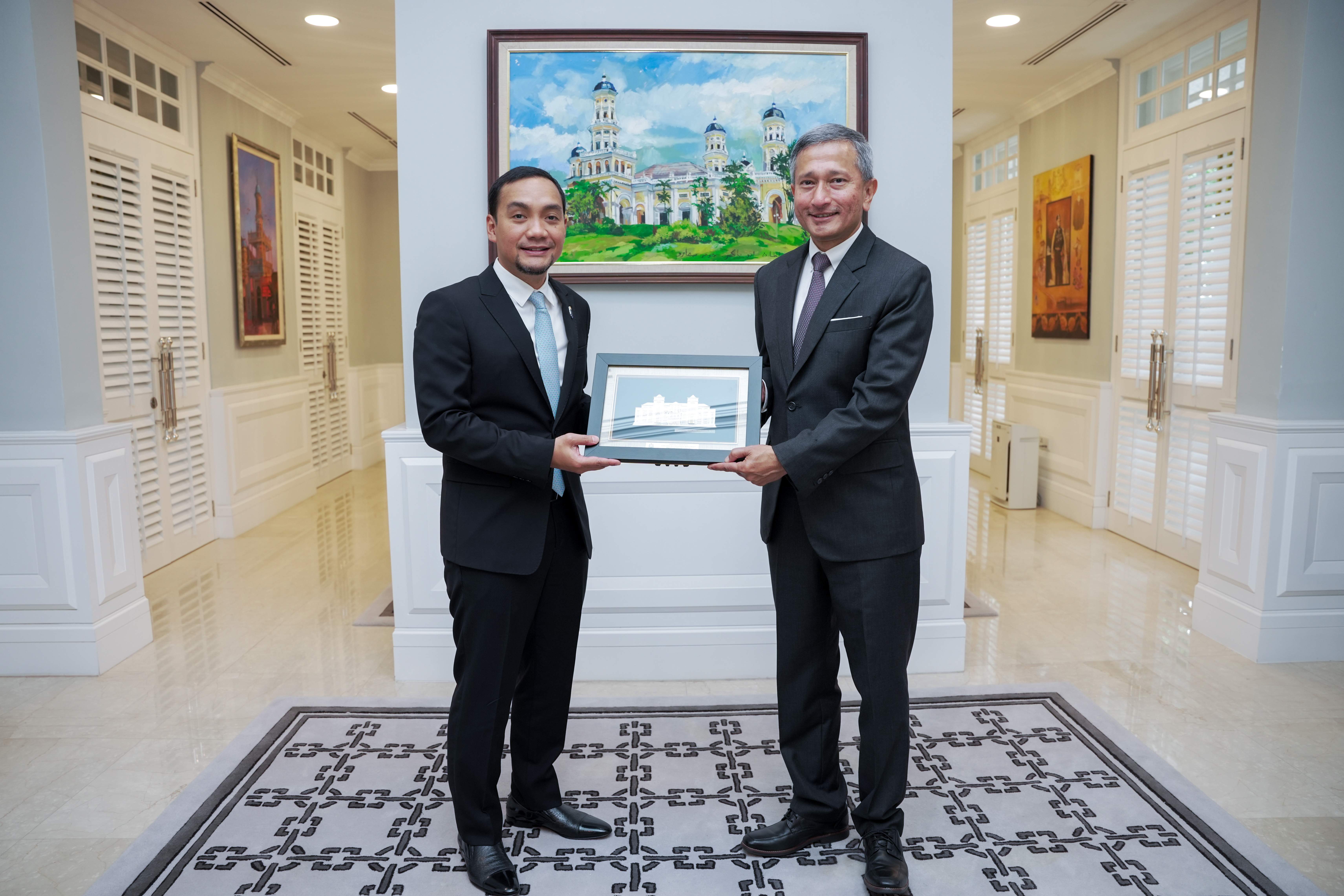 Two men in suits hold framed art beneath a painting of a building.